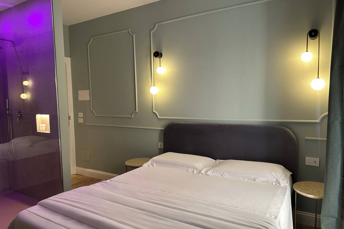 A hotel room with a large bed, two bedside tables, a mirror above the bed, and a shower area with purple lighting.