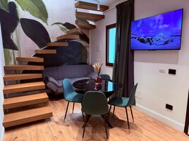 A modern living room with a staircase, a grey sofa, teal chairs, a black coffee table, and a TV displaying a snowy landscape.
