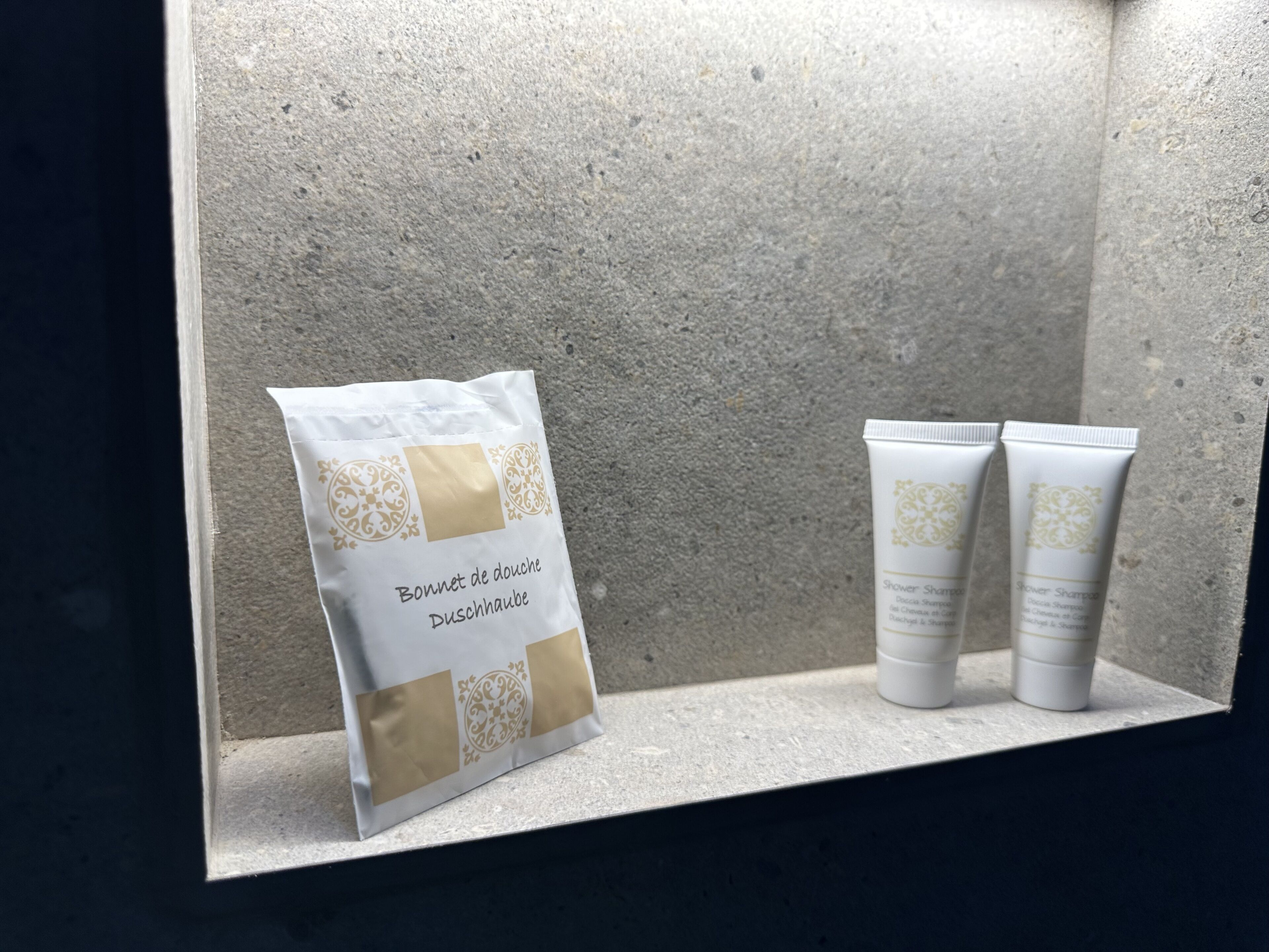 A bathroom shelf with a white and gold toiletry bag and two white tubes of shower shampoo.