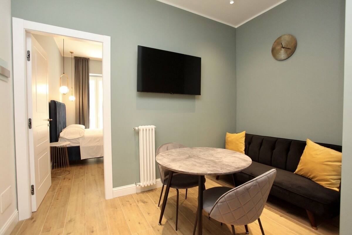A modern living room with a round table, a sofa with yellow cushions, a mounted TV, and a radiator.