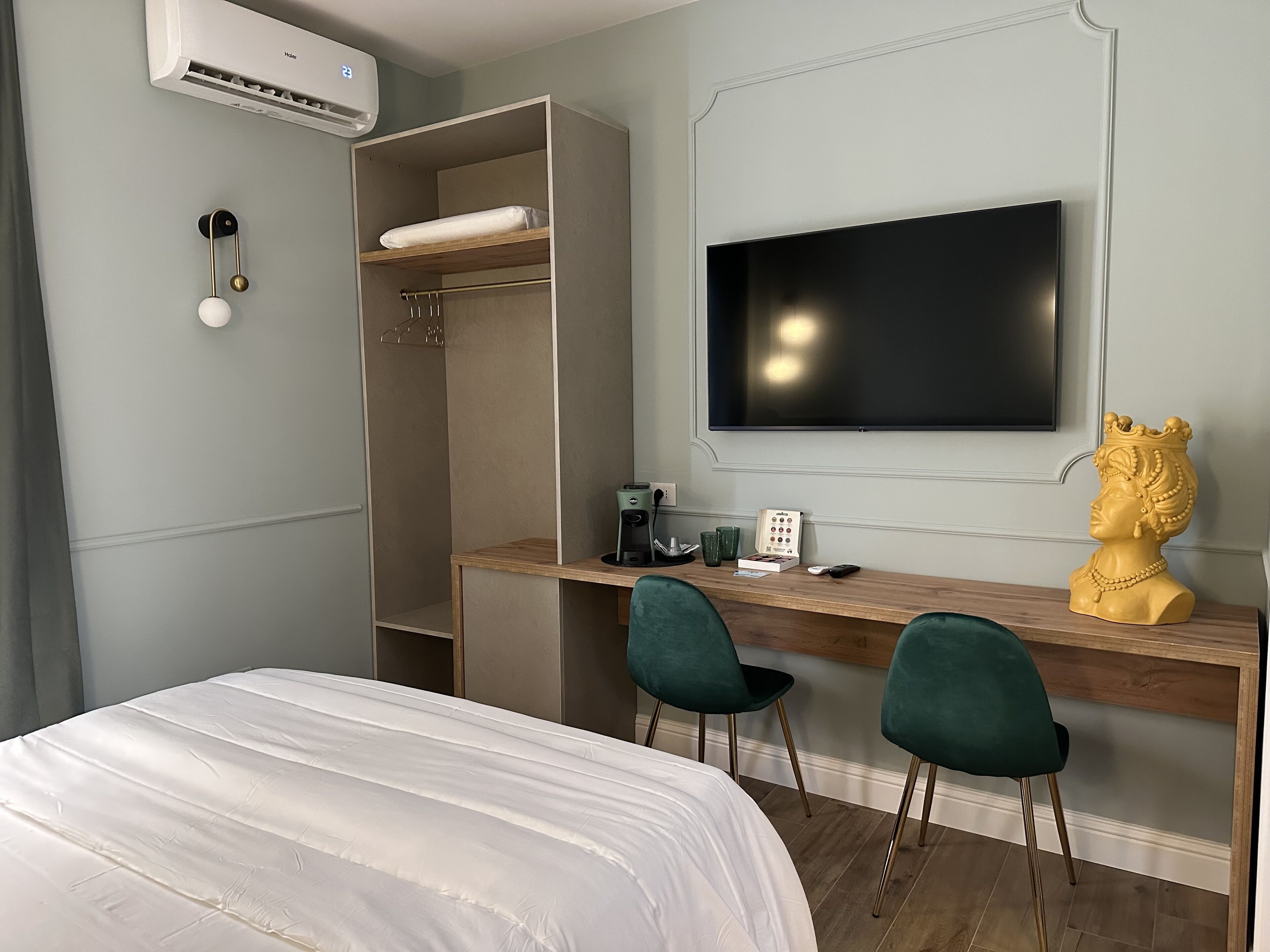 A hotel room with a bed, a desk with two chairs, a television, an air conditioner, and a decorative bust.