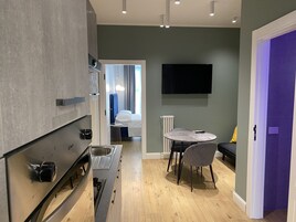 A modern kitchen with a built-in oven and sink, a dining area with a round table and chairs, and a large flat-screen TV mounted on the wall.