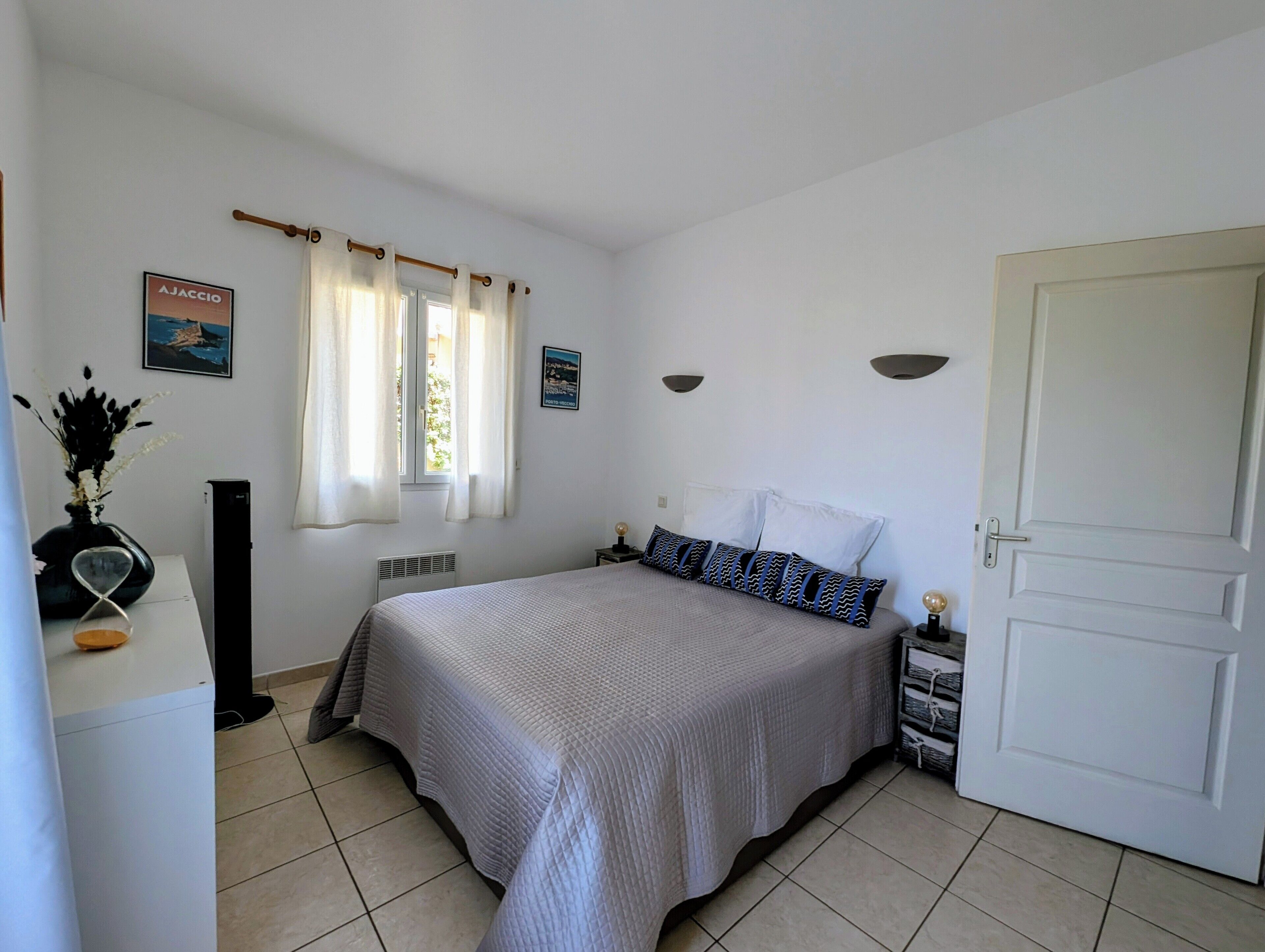 Une chambre à coucher avec un lit, une fenêtre avec des rideaux, un radiateur, une étagère fixée au mur et une porte.