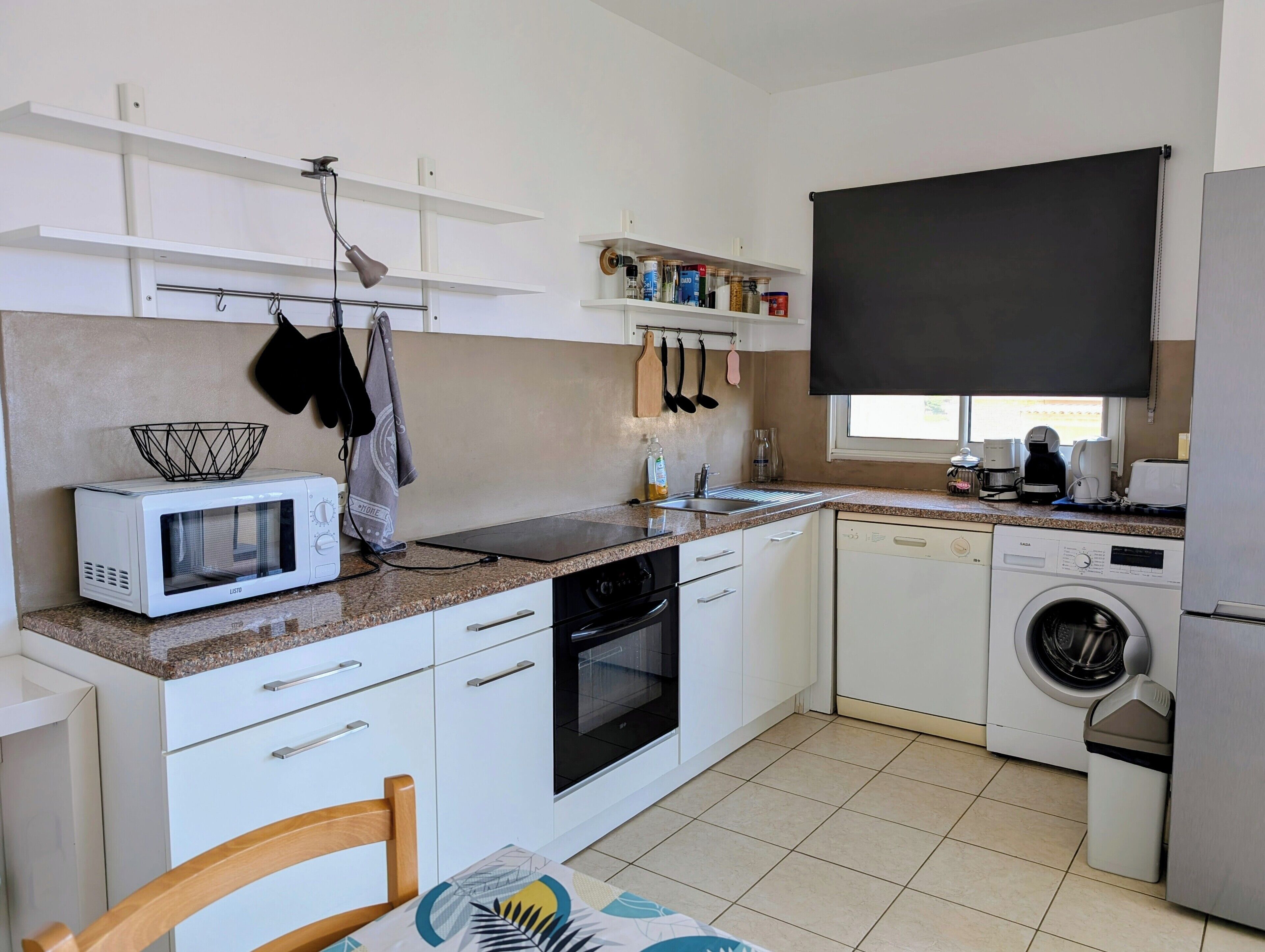 Eine kleine Küche mit weißen Schränken, einer Mikrowelle, einem Backofen, einem Geschirrspüler und einer Waschmaschine. Über der Spüle befindet sich ein Fenster, und Küchenutensilien sind an der Wand angebracht.
