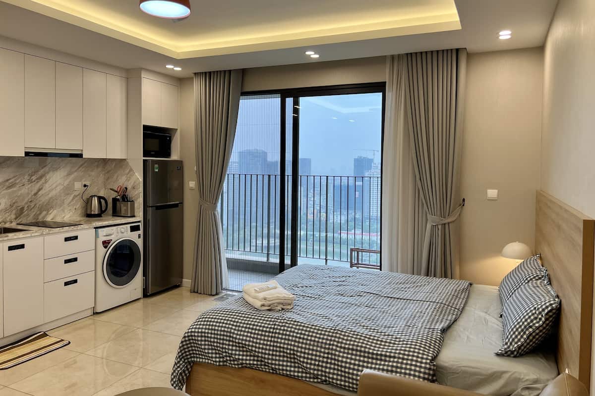 Ein modernes Hotelzimmer mit Balkon, einem Bett mit Karomuster-Bettwäsche, einer Waschmaschine und einer Küchenzeile.