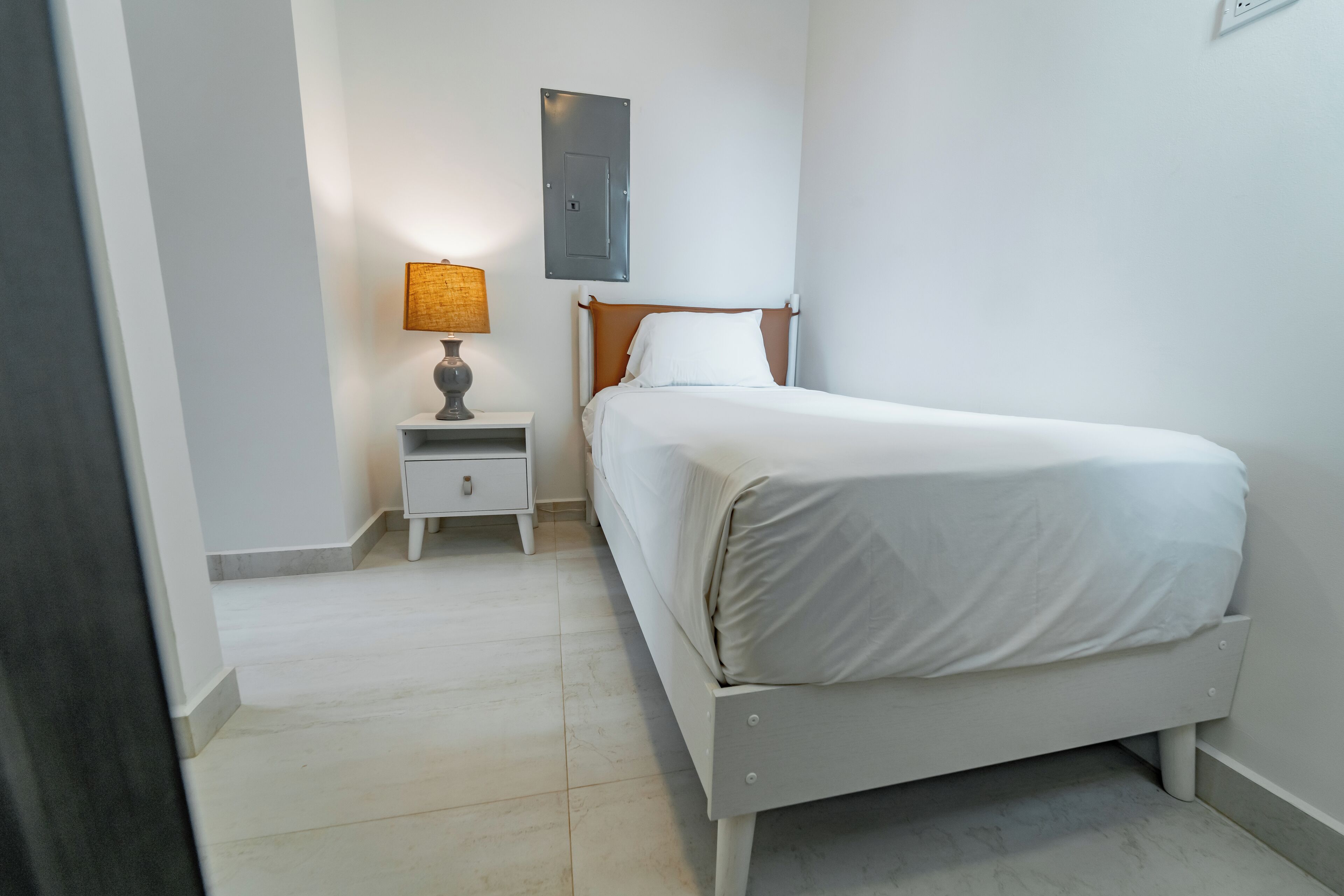 A bedroom with a bed, nightstand, lamp, and a wall-mounted electrical box.