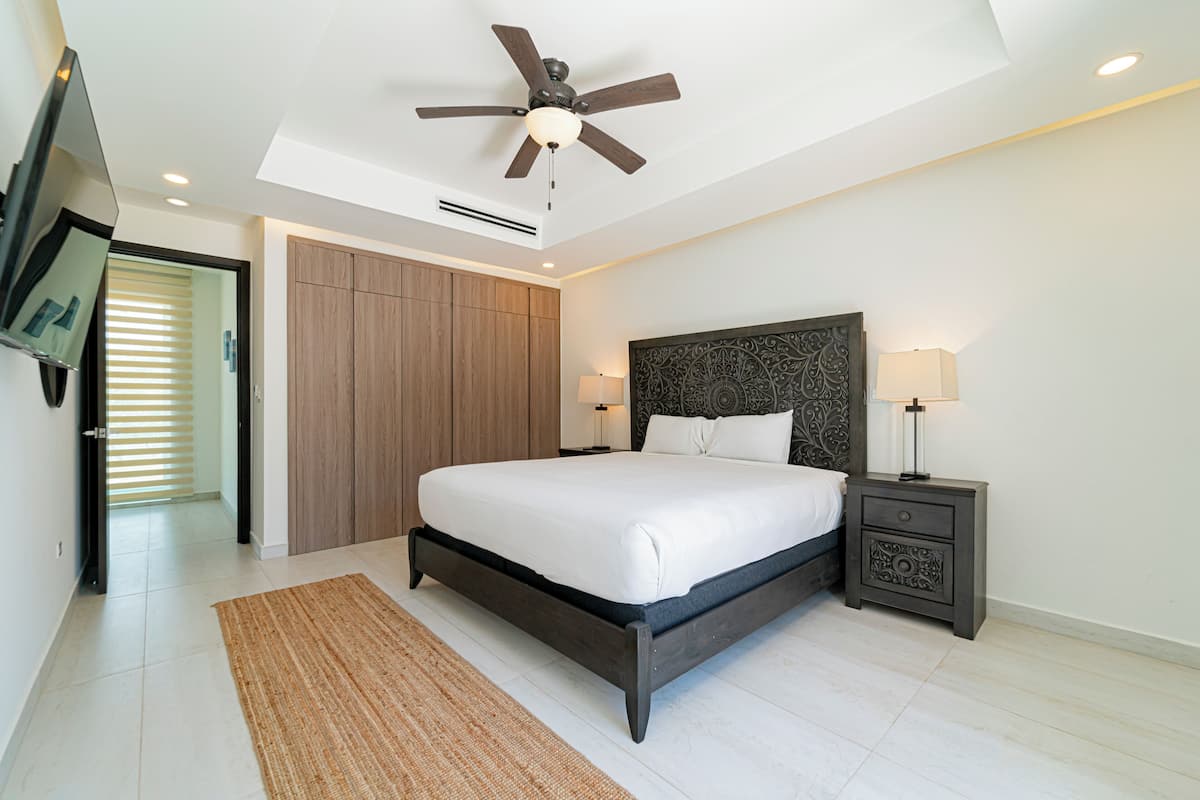 Ein Schlafzimmer mit einem Bett, einem Nachttisch, einem Deckenventilator, einem Fernseher und einer Tür zu einem anderen Raum.
