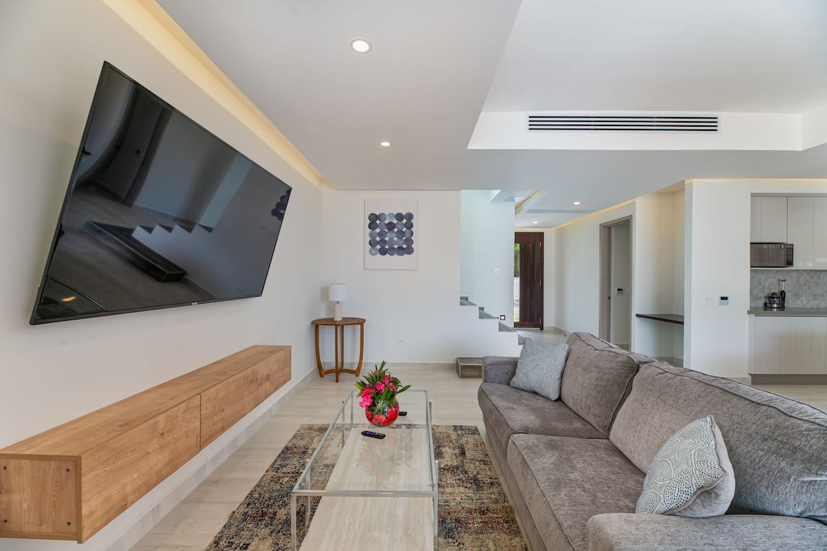 Ein modernes Wohnzimmer mit einem an der Wand befestigten Flachbildfernseher, einer Holzbank, einem Glastisch mit Blumenvase und einem grauen Sofa mit dekorativen Kissen.