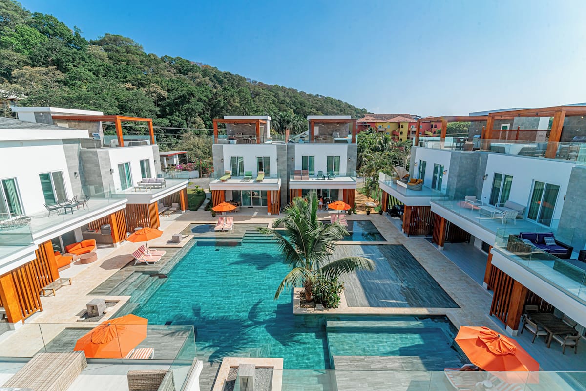 Ein modernes Hotel mit einem zentralen Schwimmbad, Sitzgelegenheiten im Freien und Blick auf einen bewaldeten Hügel.