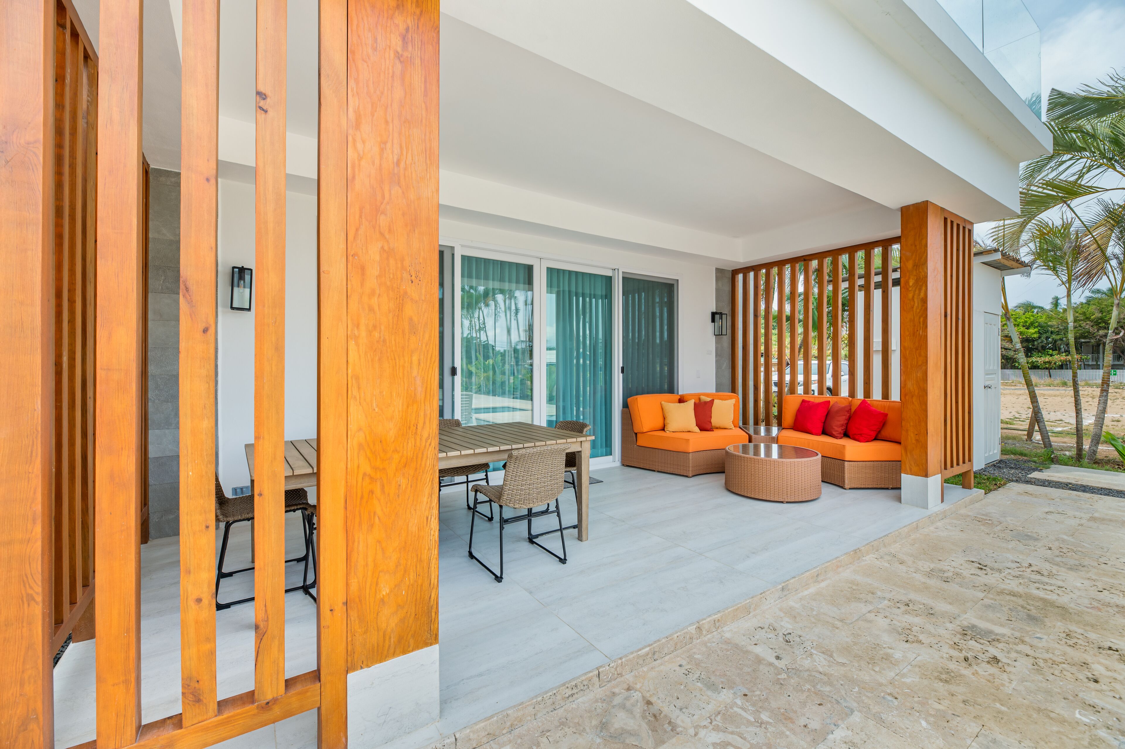 A modern outdoor seating area with a wooden table and chairs, a sofa with orange cushions, and a wicker coffee table.