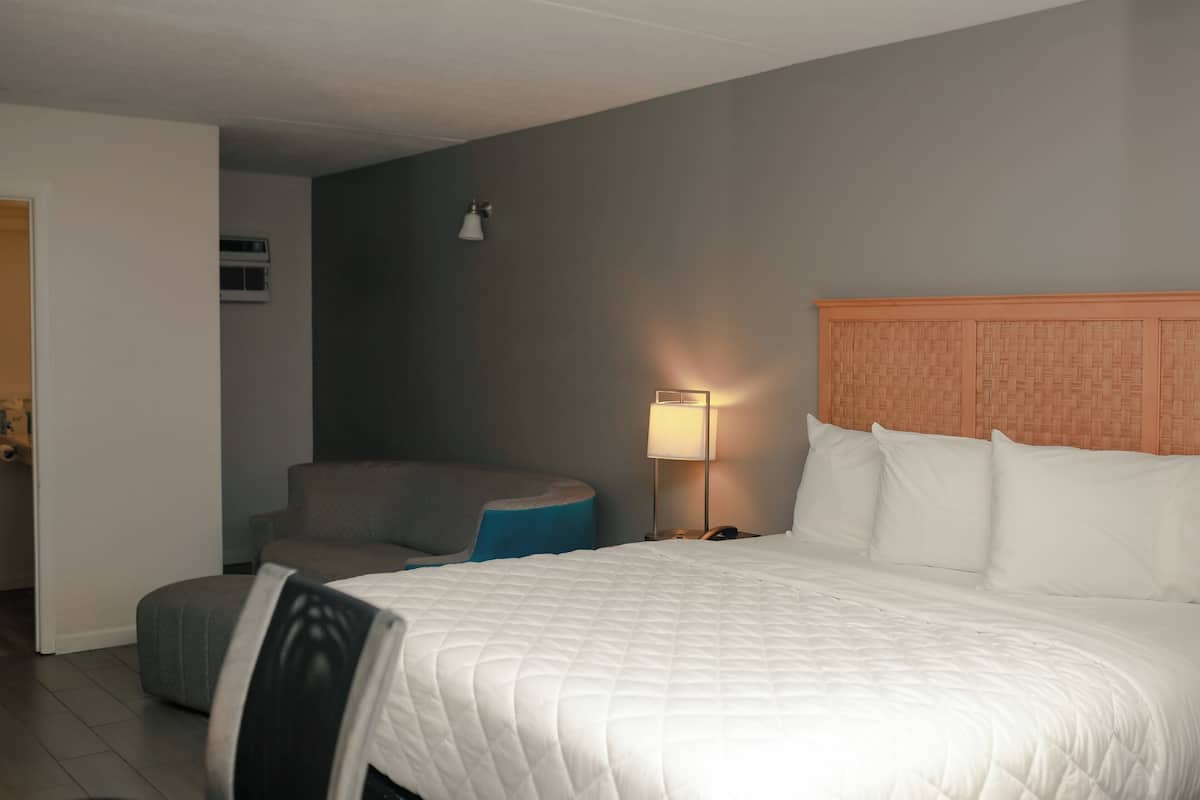 A hotel room with a bed, a nightstand with a lamp, a chair, and a small ottoman.