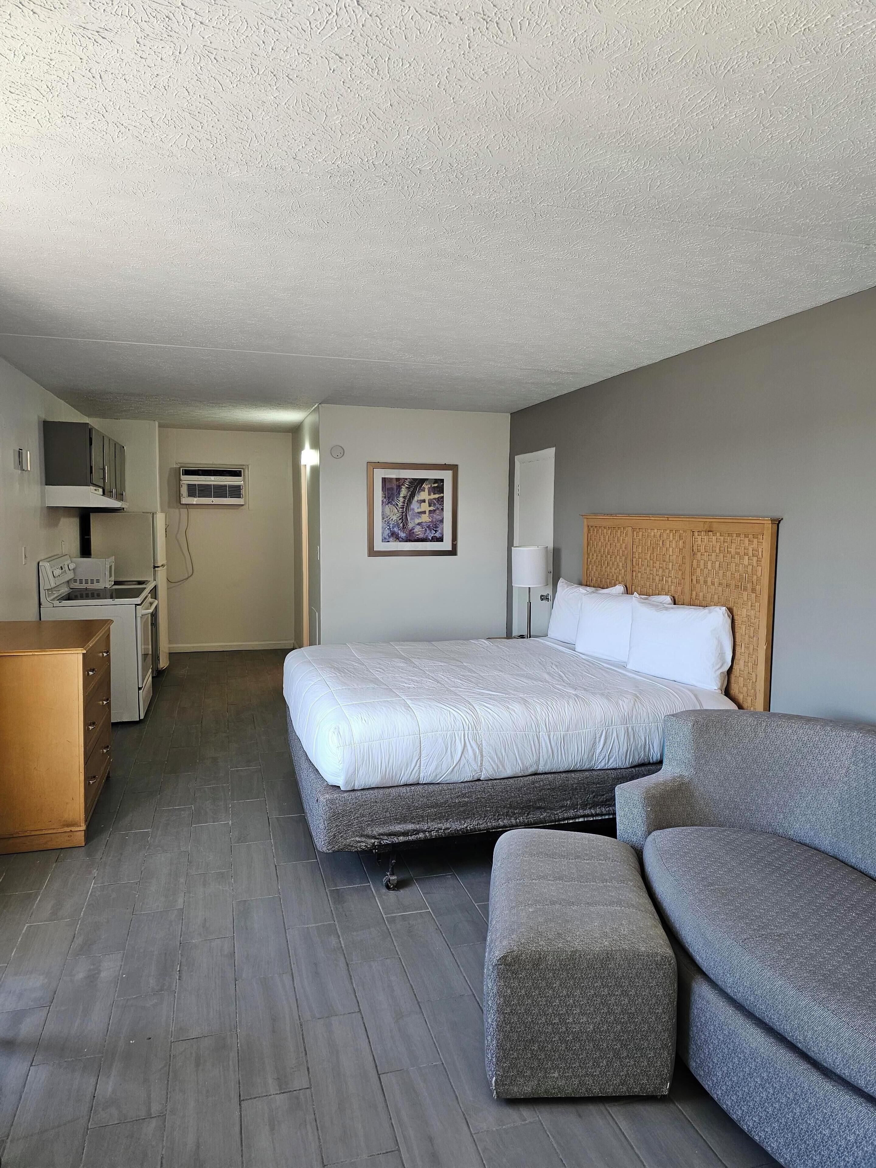 A hotel room with a bed, a sofa, a wooden dresser, a microwave, and an air conditioning unit.