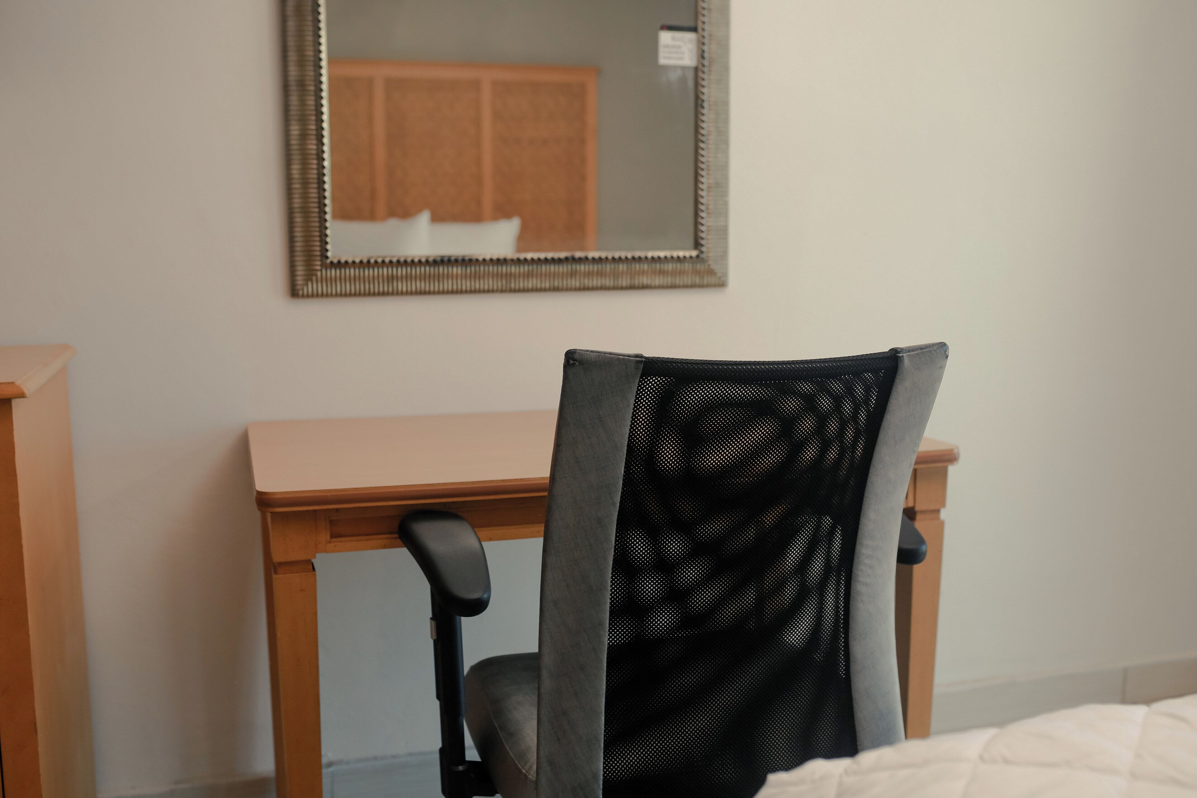 A hotel room with a wooden desk, a black mesh chair, and a mirror on the wall.