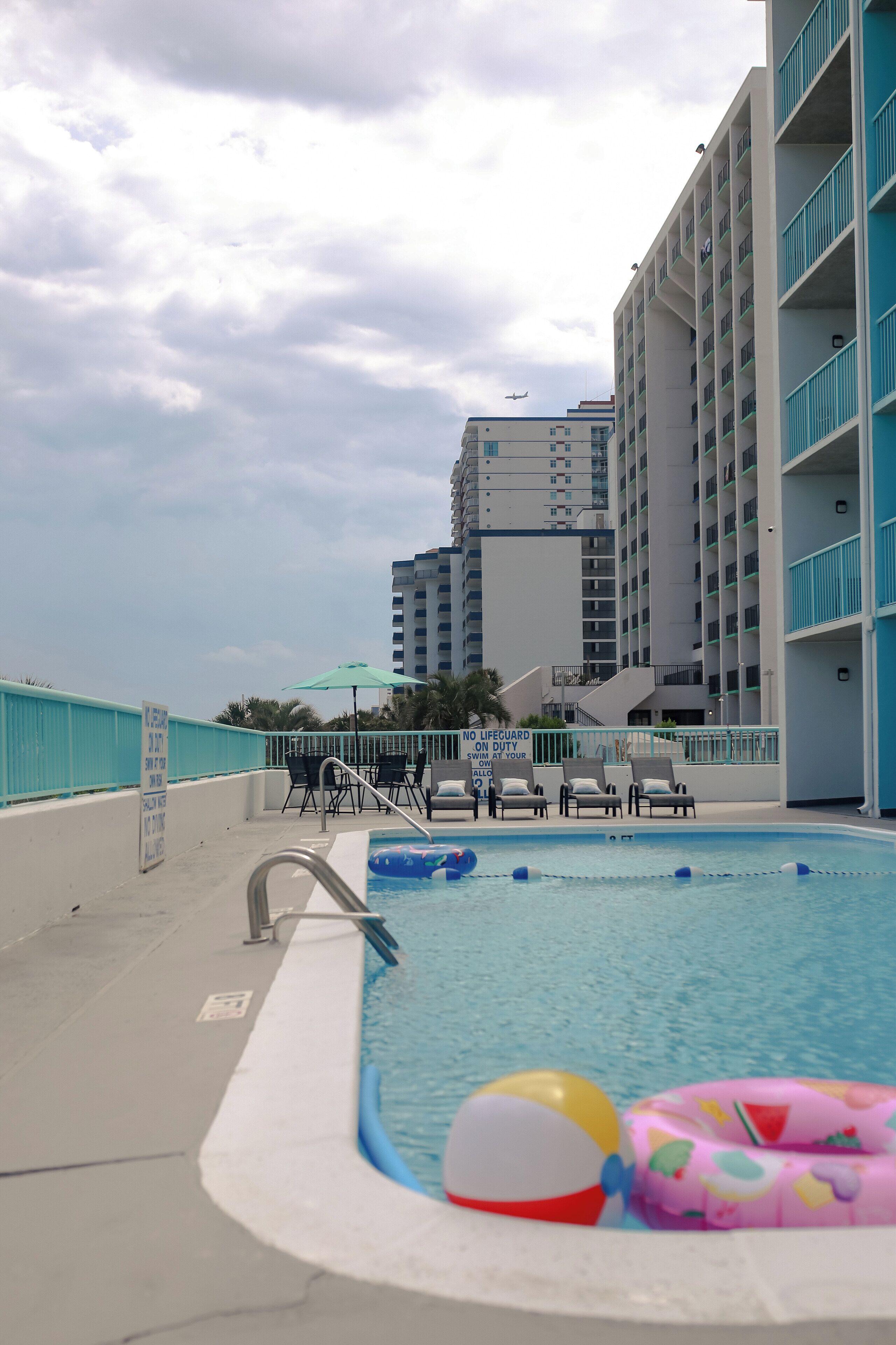 A swimming pool with lounge chairs and inflatable toys, surrounded by a high-rise building with balconies.