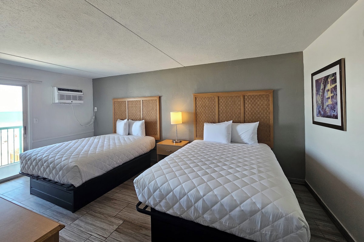 A hotel room with two beds, a bedside table, a lamp, a framed picture on the wall, and an air conditioning unit.