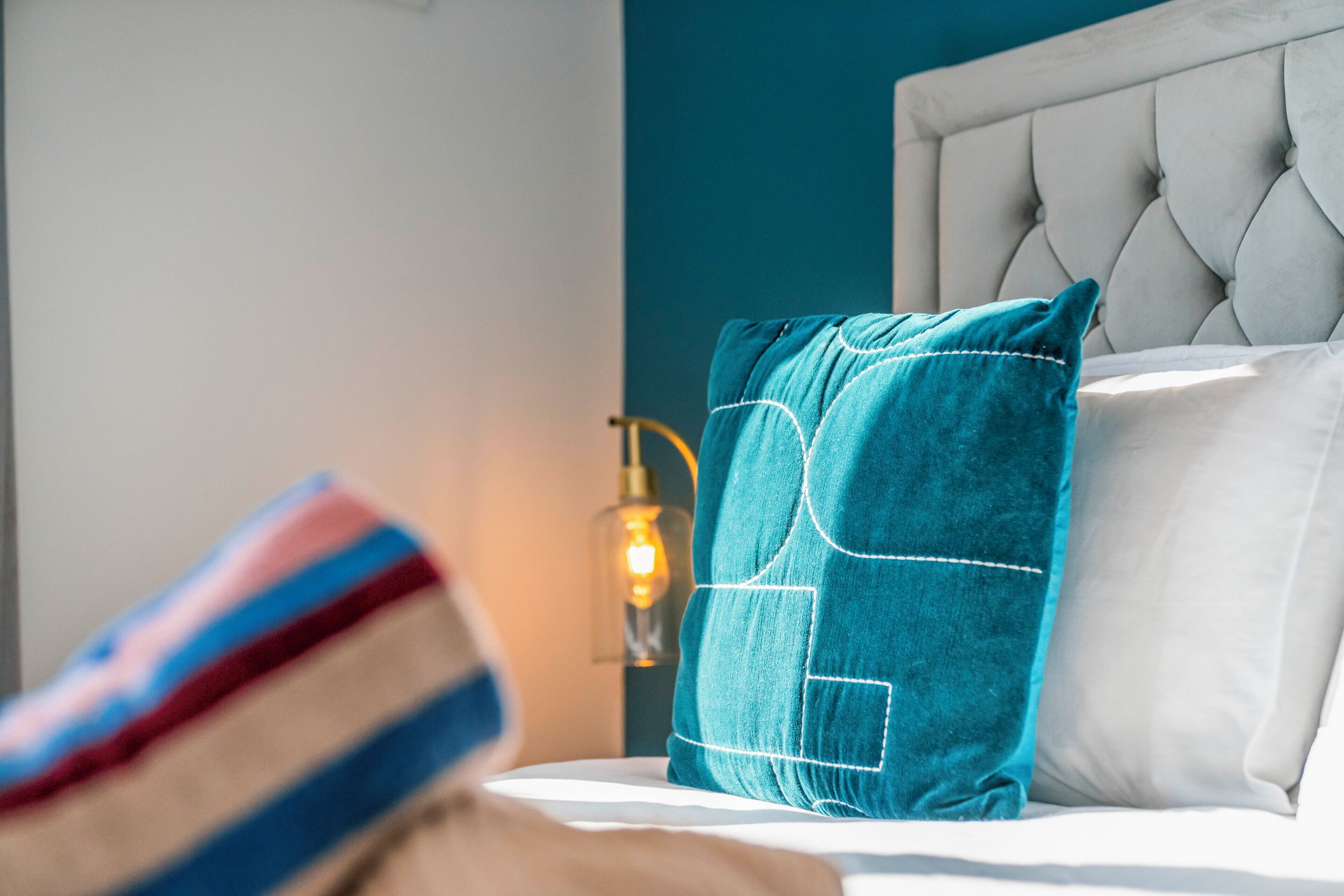 A bed with a tufted headboard, a teal pillow, and a white pillow. A lamp with a glass shade and a visible light bulb is on the nightstand.