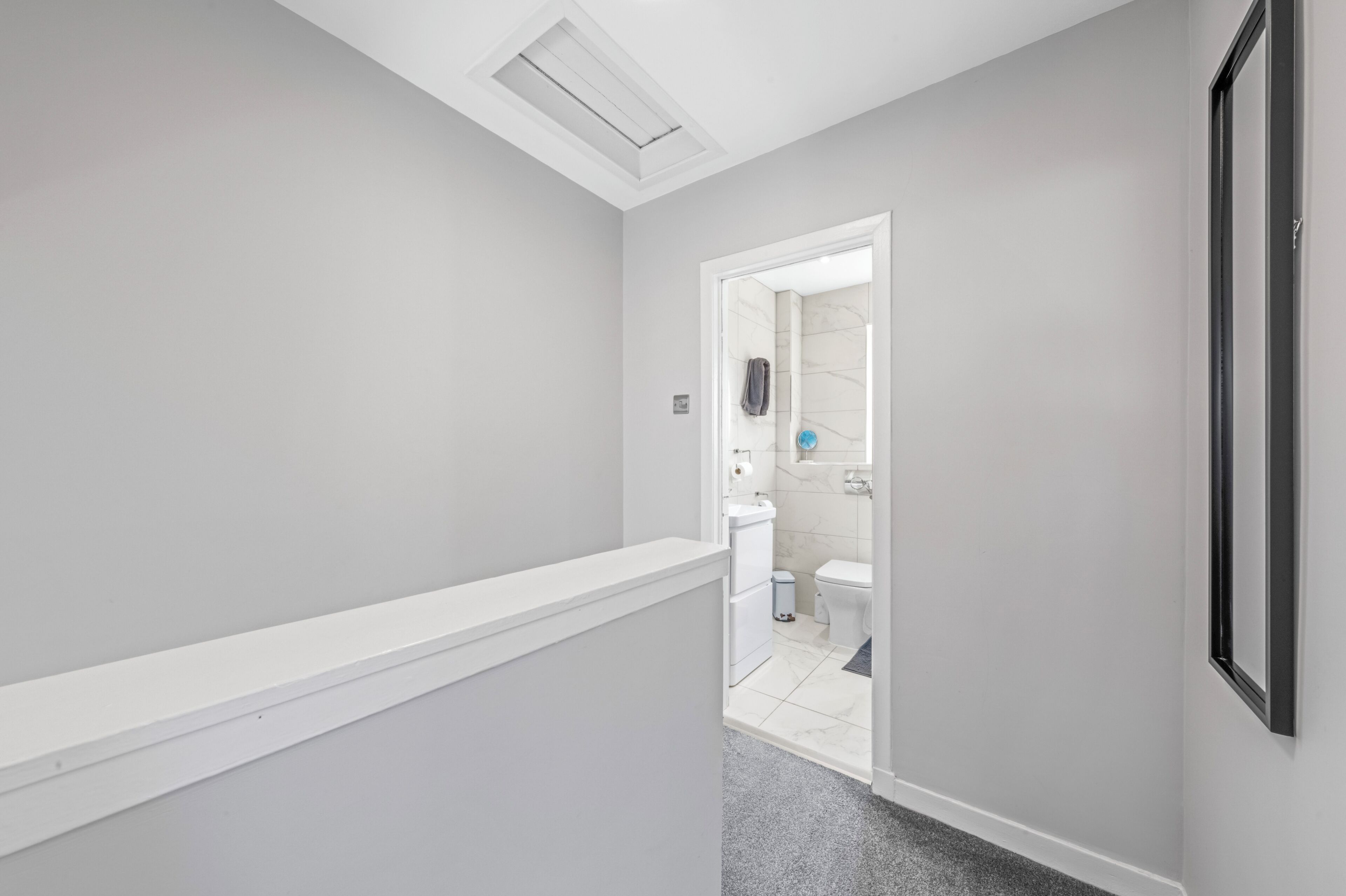 A narrow hallway with a white ceiling and a door leading to a bathroom with a toilet and a sink.