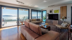 Ein modernes Wohnzimmer mit einer großen Couch, einem Couchtisch und einem Fernseher. Große Fenster bieten Blick auf das Meer.