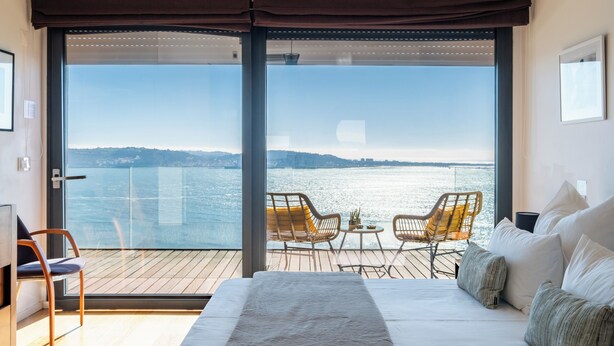 Ein Schlafzimmer mit großem Fenster und Blick auf ein Gewässer, einem Bett mit weißer Bettwäsche, zwei Korbstühlen und einem kleinen Tisch.