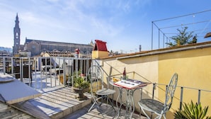 Una terraza en la azotea con sillas de metal, una mesa, plantas en macetas y vistas de un edificio histórico con una torre de reloj.