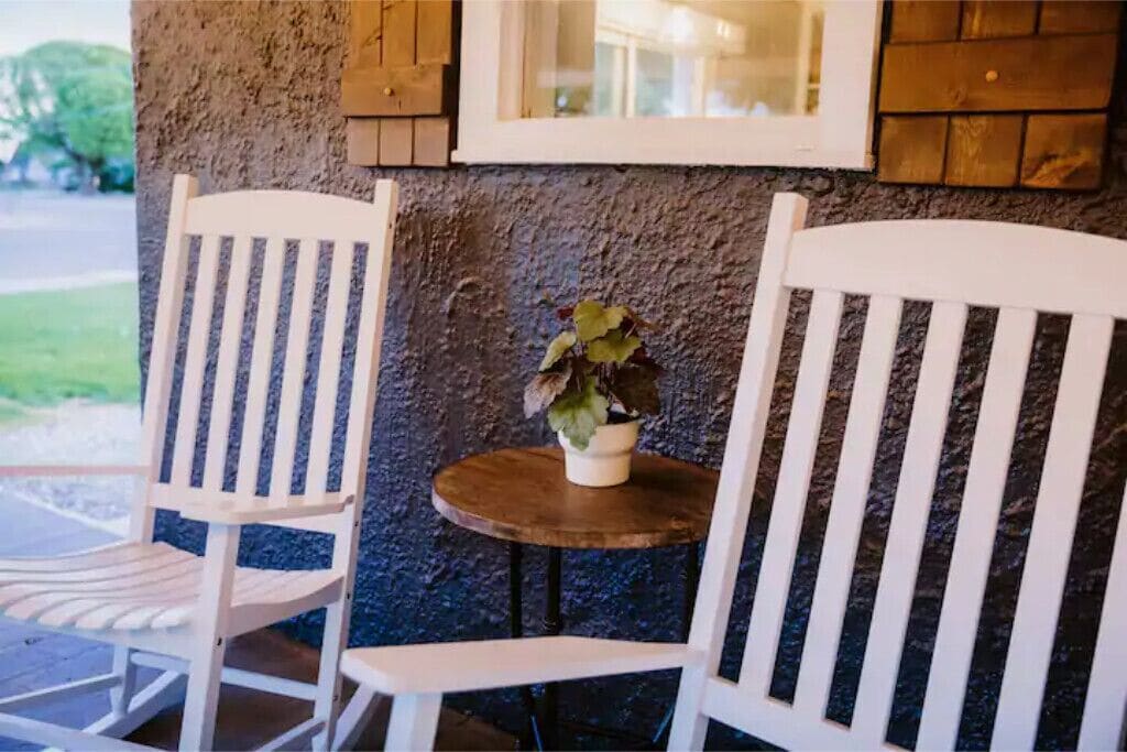 Deux chaises blanches et une petite table ronde, sur laquelle est posée une plante en pot, se trouvent sur une véranda dont le mur présente une texture particulière et qui est agrémentée d’éléments en bois.