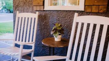 Deux chaises blanches et une petite table ronde, sur laquelle est posée une plante en pot, se trouvent sur une véranda dont le mur présente une texture particulière et qui est agrémentée d’éléments en bois.