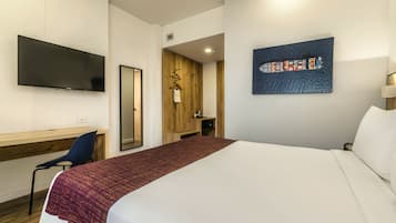 Quarto de hotel com cama, escrivaninha, cadeira, televisão e um quadro na parede.
