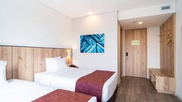 A hotel room with two beds, a wooden headboard, a wall-mounted artwork, a bench, and a door with a sign.