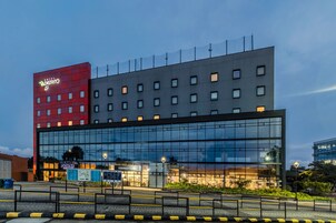 Un hotel moderno con fachada roja y gris, amplias ventanas de vidrio y un restaurante visible llamado "Plaza de los Frescos".