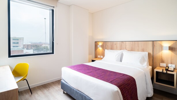 A hotel room with a bed, a desk, a chair, a window, and a nightstand with a lamp. - Hotel Distrito ZF By OxoHotel (Bogotá)