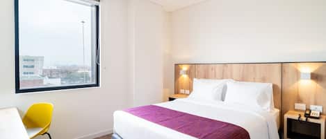 A hotel room with a bed, a desk, a chair, a window, and a nightstand with a lamp.