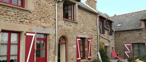 A stone building with red shutters and a small garden area with a chair and wheelbarrow.