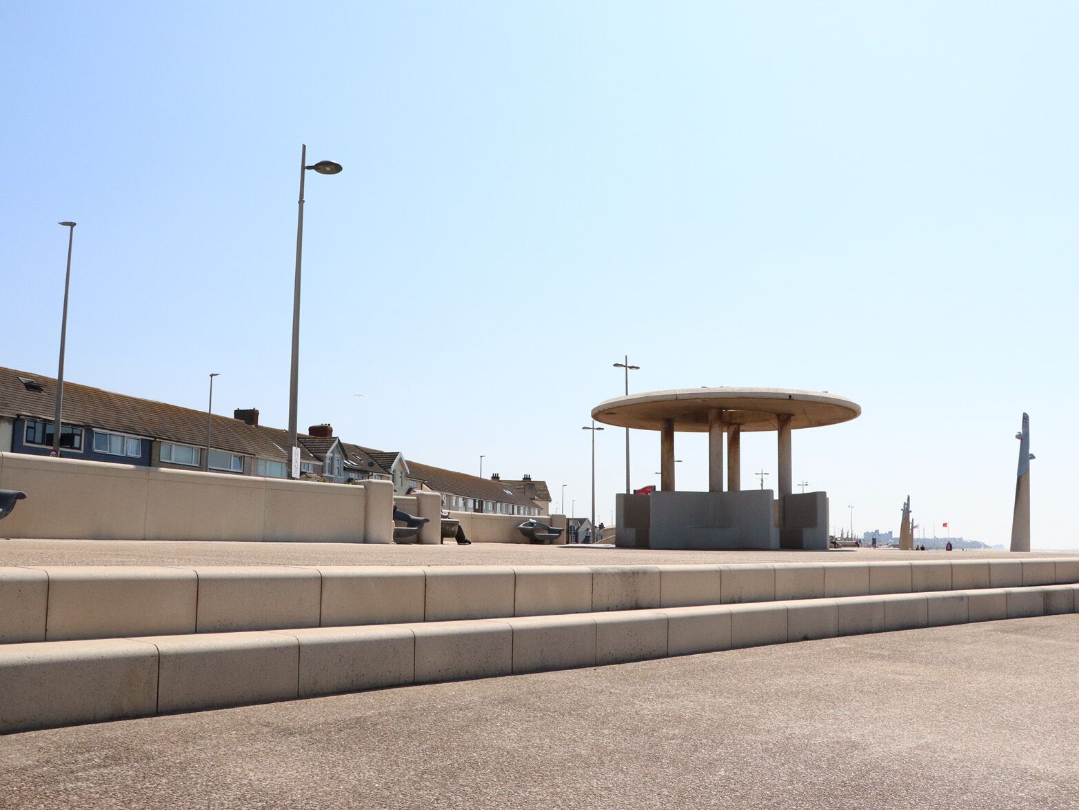 A seaside promenade with a circular monument, steps, and residential buildings in the background.