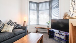 Een woonkamer met een bank, een houten salontafel, een tv-meubel met een flatscreen televisie en een raam met jaloezieën.