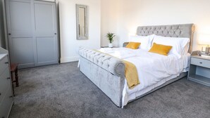 Een slaapkamer met een grijs gewatteerd hoofdeinde, wit beddengoed en een mosterdgele plaid.