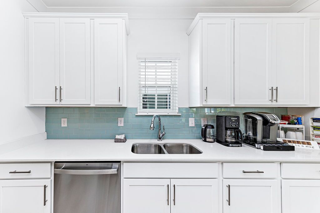 A kitchen with white cabinets, a stainless steel sink, a window with blinds, and a coffee maker.