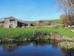 Une maison en pierre, avec un étang au premier plan, entourée d’une pelouse et d’arbres.