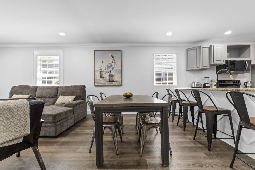 A modern kitchen with white cabinets, a dining area with a wooden table and metal chairs, and a gray sofa with a patterned throw.