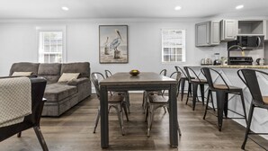 Une cuisine moderne avec des meubles blancs, un coin repas avec une table en bois et des chaises en métal, et un canapé gris agrémenté d’un plaid à motifs.