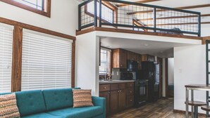Een ruime loft-achtige woonruimte met een hoge nok, houten balken, een keuken met donkere kastjes en een blauwe bank met opdrukkeussens.