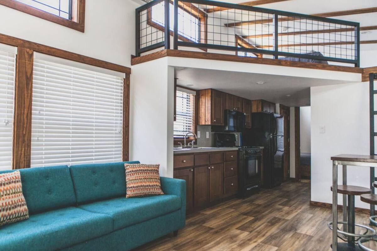 A spacious loft-style living area with a high ceiling, wooden beams, a kitchen with dark cabinets, and a blue sofa with patterned cushions.