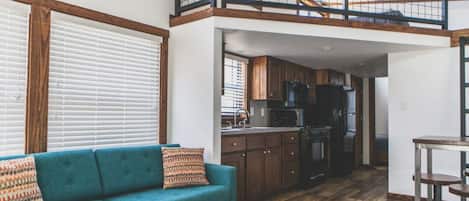A spacious loft-style living area with a high ceiling, wooden beams, a kitchen with dark cabinets, and a blue sofa with patterned cushions.