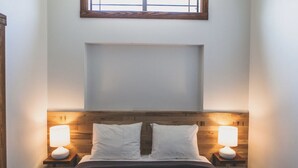 Een slaapkamer met een houten hoofdbord, twee bedlampen, een groot raam boven het bed, en een lege muur boven het hoofdbord.