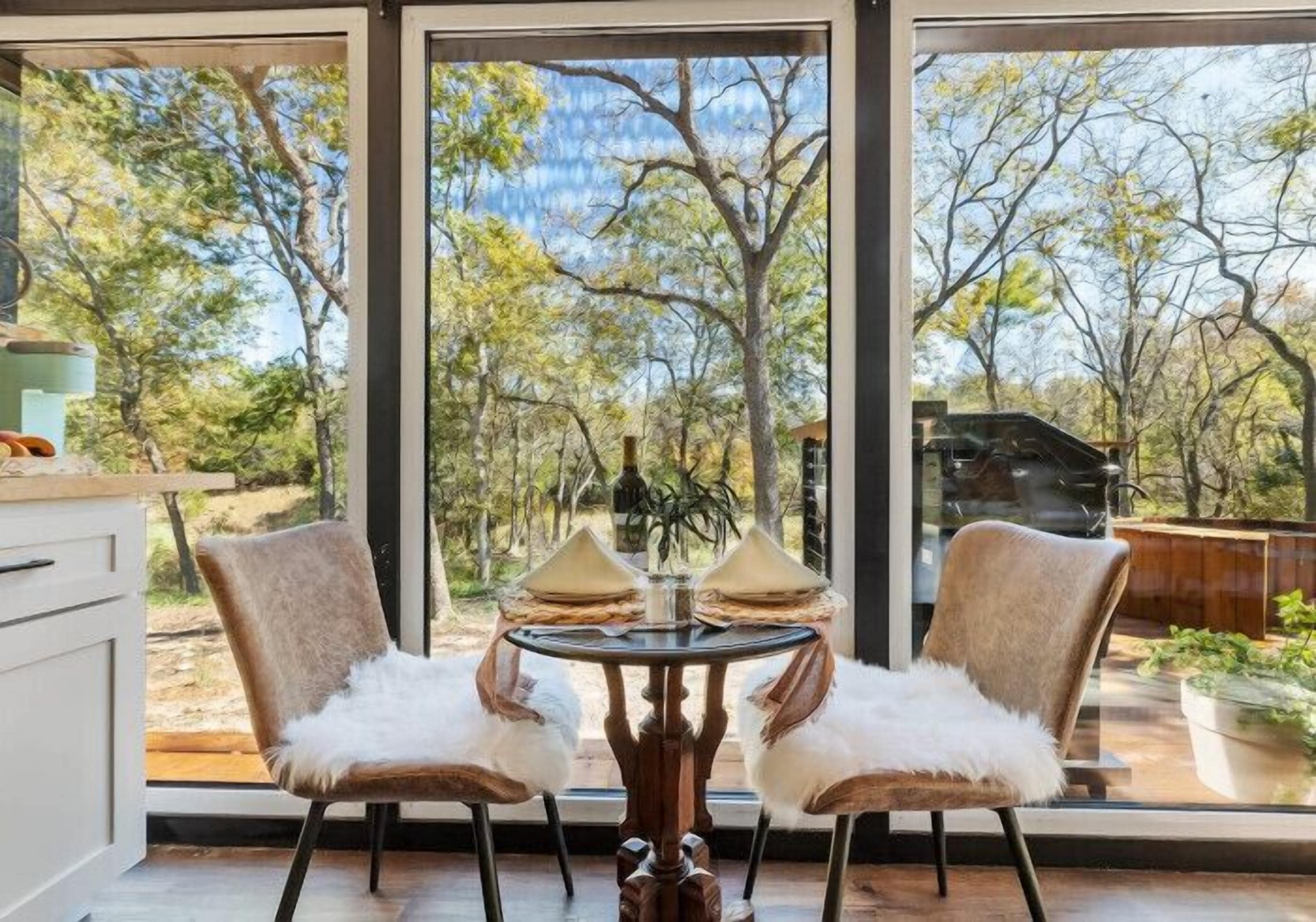 A modern kitchen with a large window, two chairs with white fur throws, and a round table with two decorative cones.