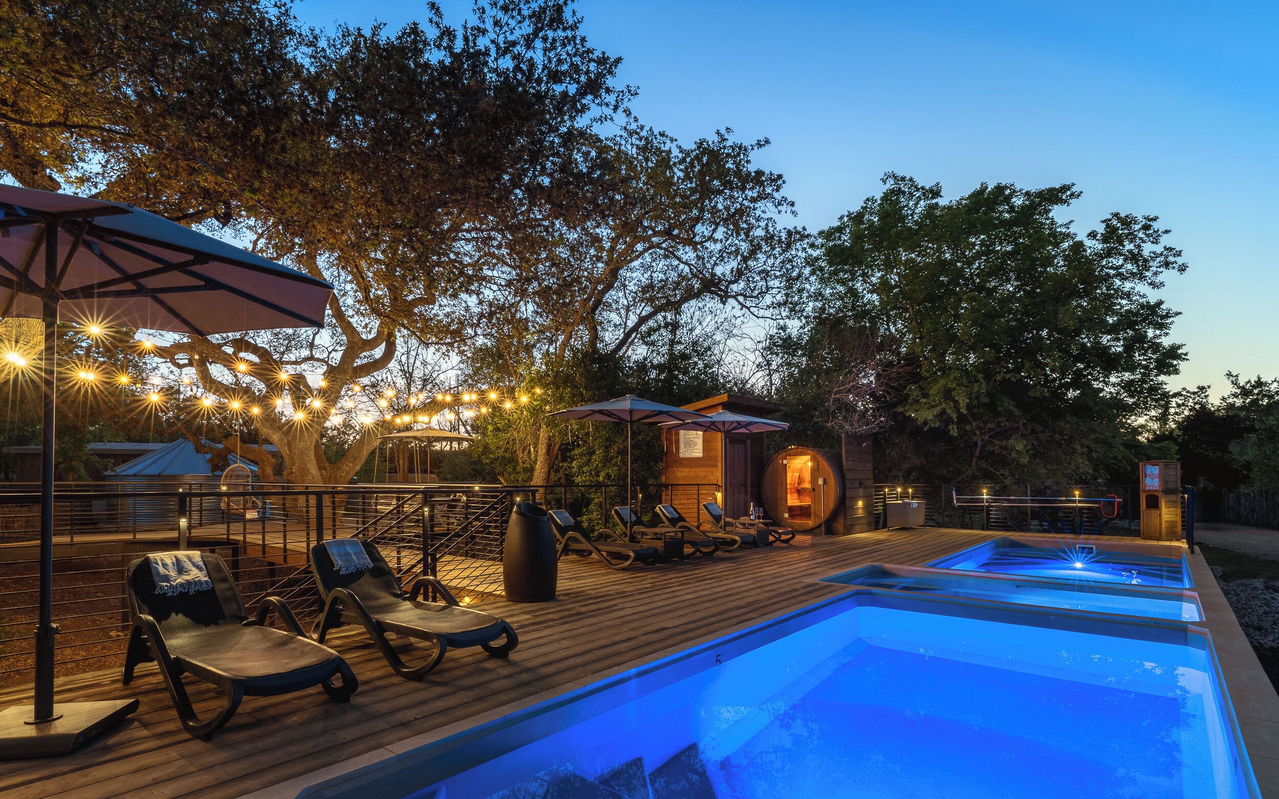 A pool area with lounge chairs and umbrellas, surrounded by trees and a wooden deck.