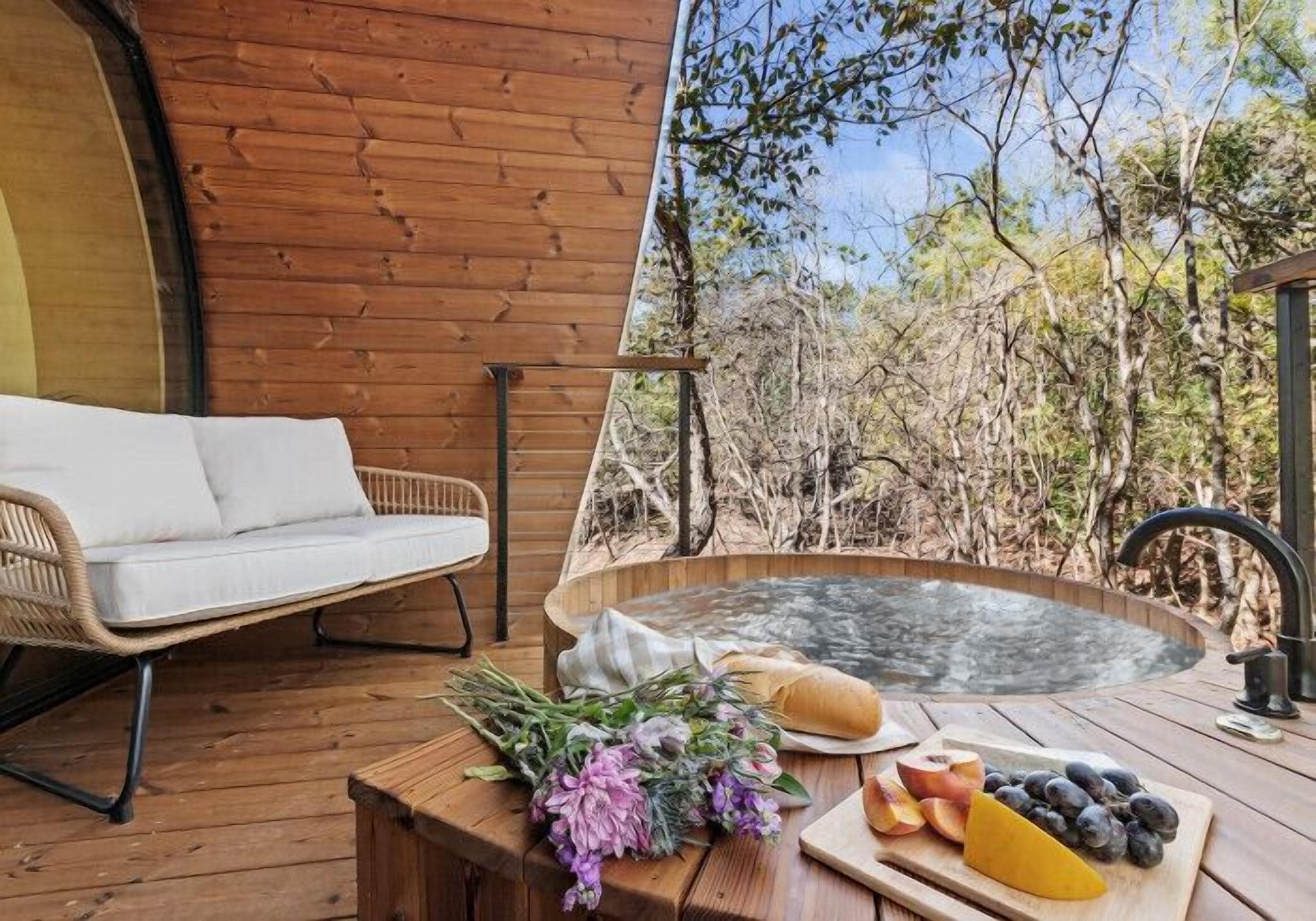 A wooden deck with a wicker sofa, a hot tub, and a table with fruits and flowers.