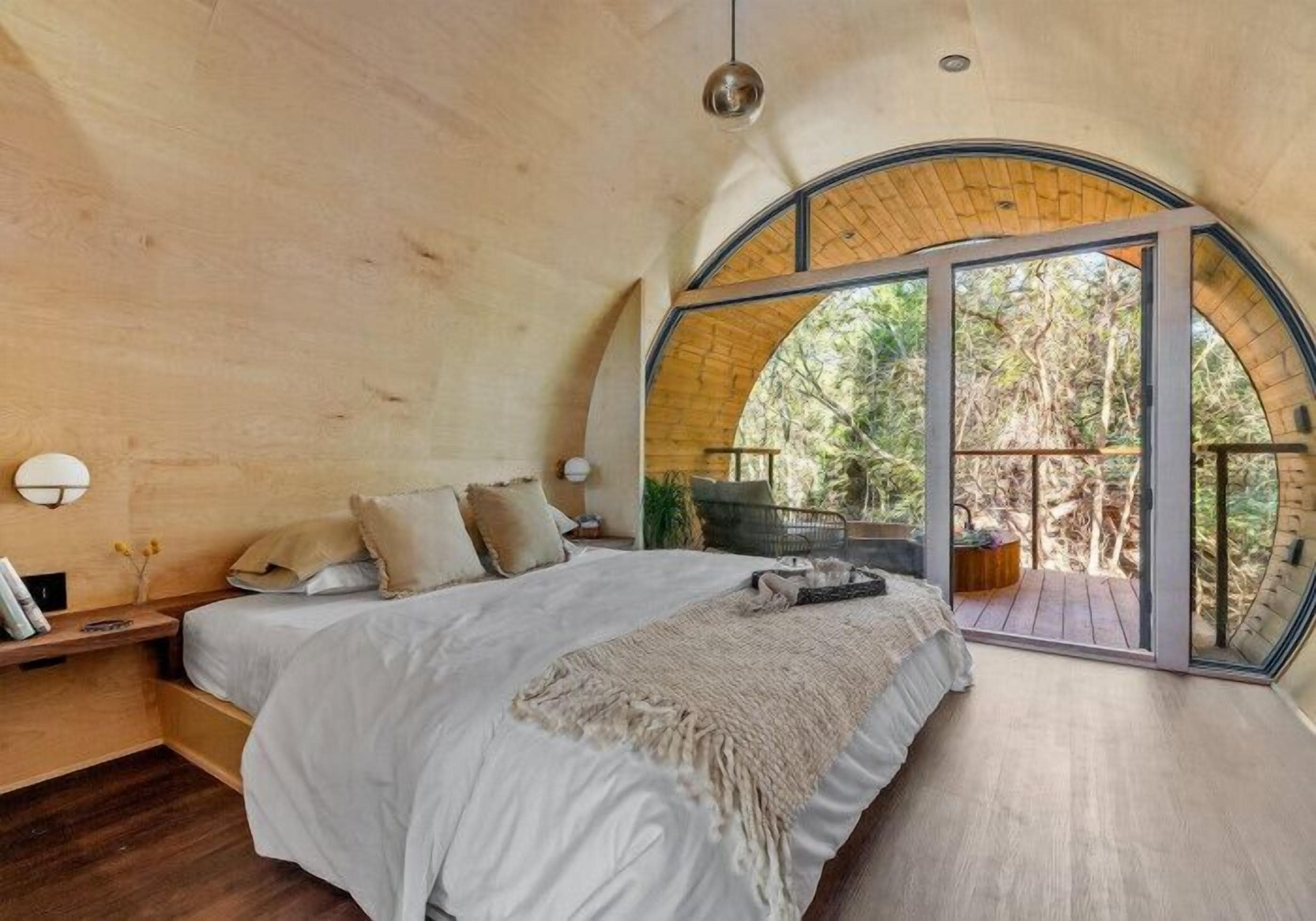 A bedroom with a large bed, a wooden headboard, a nightstand, a bedside table, and a large arched window offering a view of the outdoors.