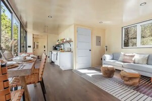 A spacious living area with a dining table set for four, a sofa, and a large window providing a view of the outdoors.