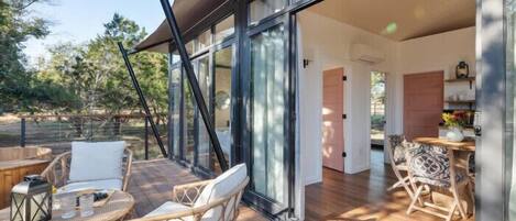 Un patio moderno al aire libre con mobiliario de mimbre y un ventanal de vidrio que da a una habitación con piso de madera y zona de comedor.