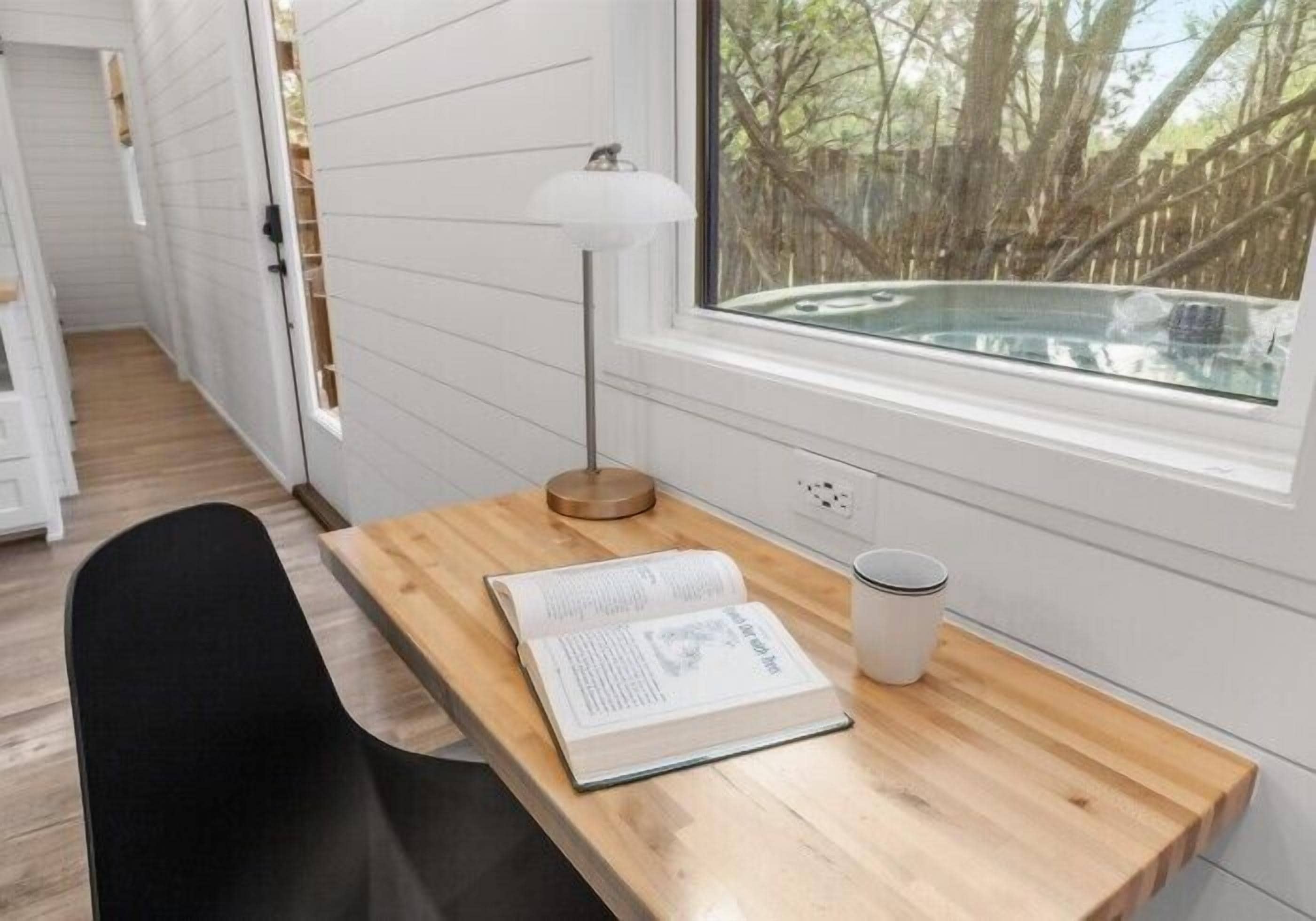Une pièce avec un bureau en bois, un livre, une lampe et une fenêtre donnant sur des arbres et un jacuzzi.